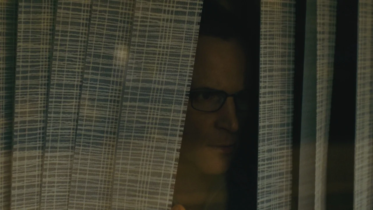 Mommy (2014) movie still: closeup — A man's face appears through a vertical gap in textured grey window blinds. He looks outward from the darkness…