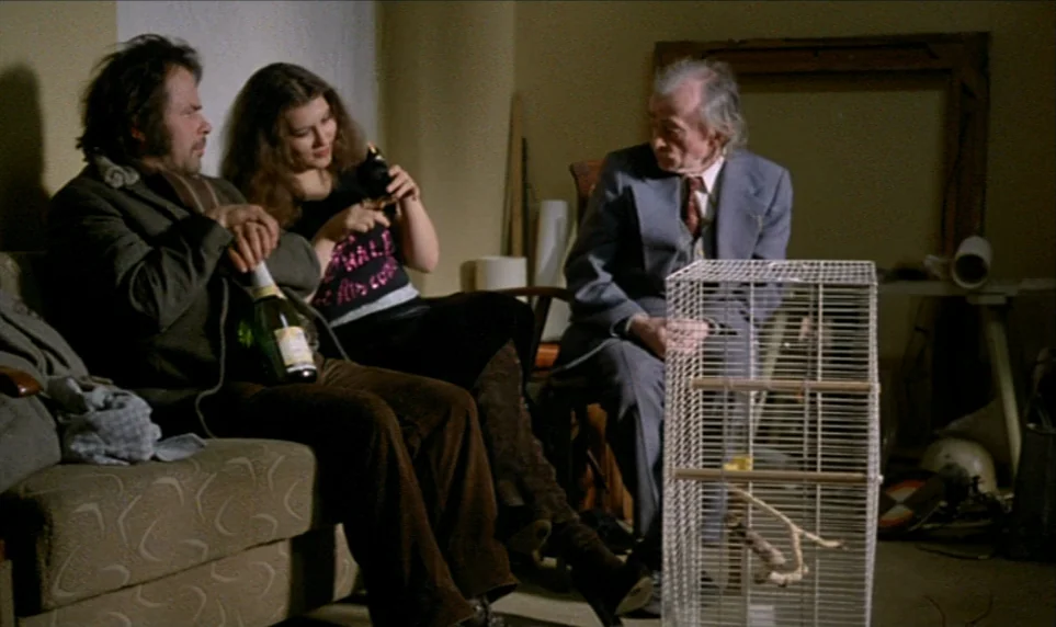Stroszek (1977) movie still: medium — A dimly lit room with plain walls and scattered items like a birdcage. Three people sit on a sofa and chair in…