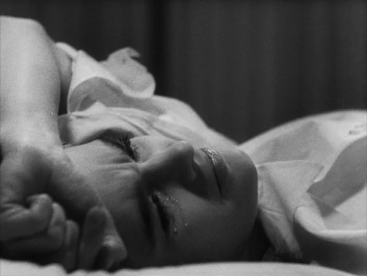 Thirst (1949) movie still: closeup — A close-up view of a person lying on white fabric in a dim space. The subject rests on their side with one arm…