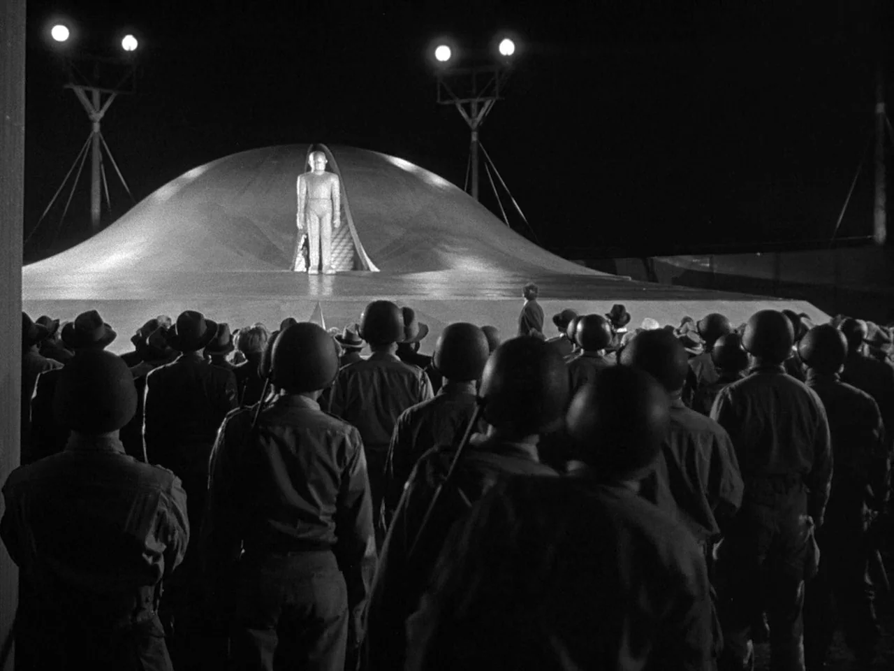 The Day the Earth Stood Still (1951) movie still: wide — A crowd of soldiers in combat helmets faces a massive, smooth, rounded spaceship. A…