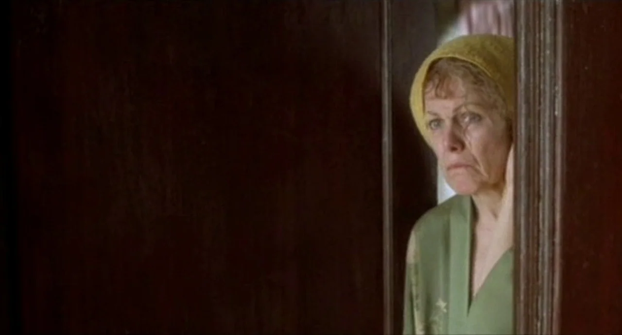 The Annihilation of Fish (1999) movie still: medium — A woman peeks out from the side of a dark wooden door frame. She is wearing a yellow head wrap…