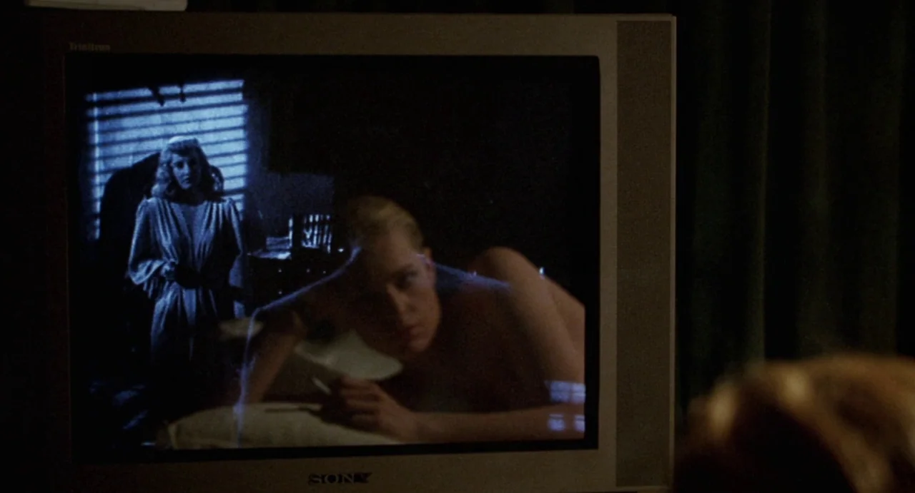 Femme Fatale (2002) movie still: closeup — A dark room contains a Sony Trinitron television displaying a grainy video image. The screen shows two…
