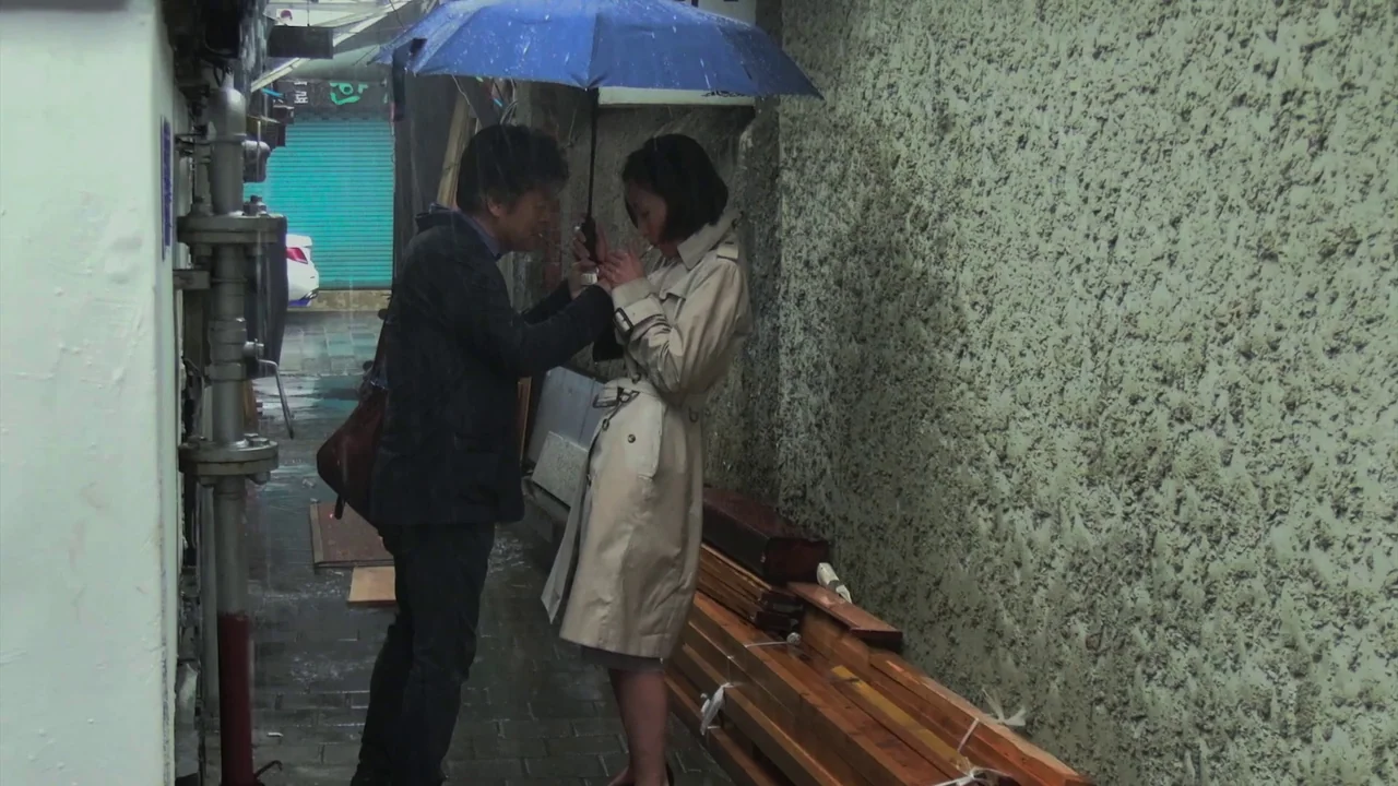 In Front Of Your Face (2021) movie still: fullbody — A narrow stone alleyway in a city during light rain. A man holds a blue umbrella over a woman…