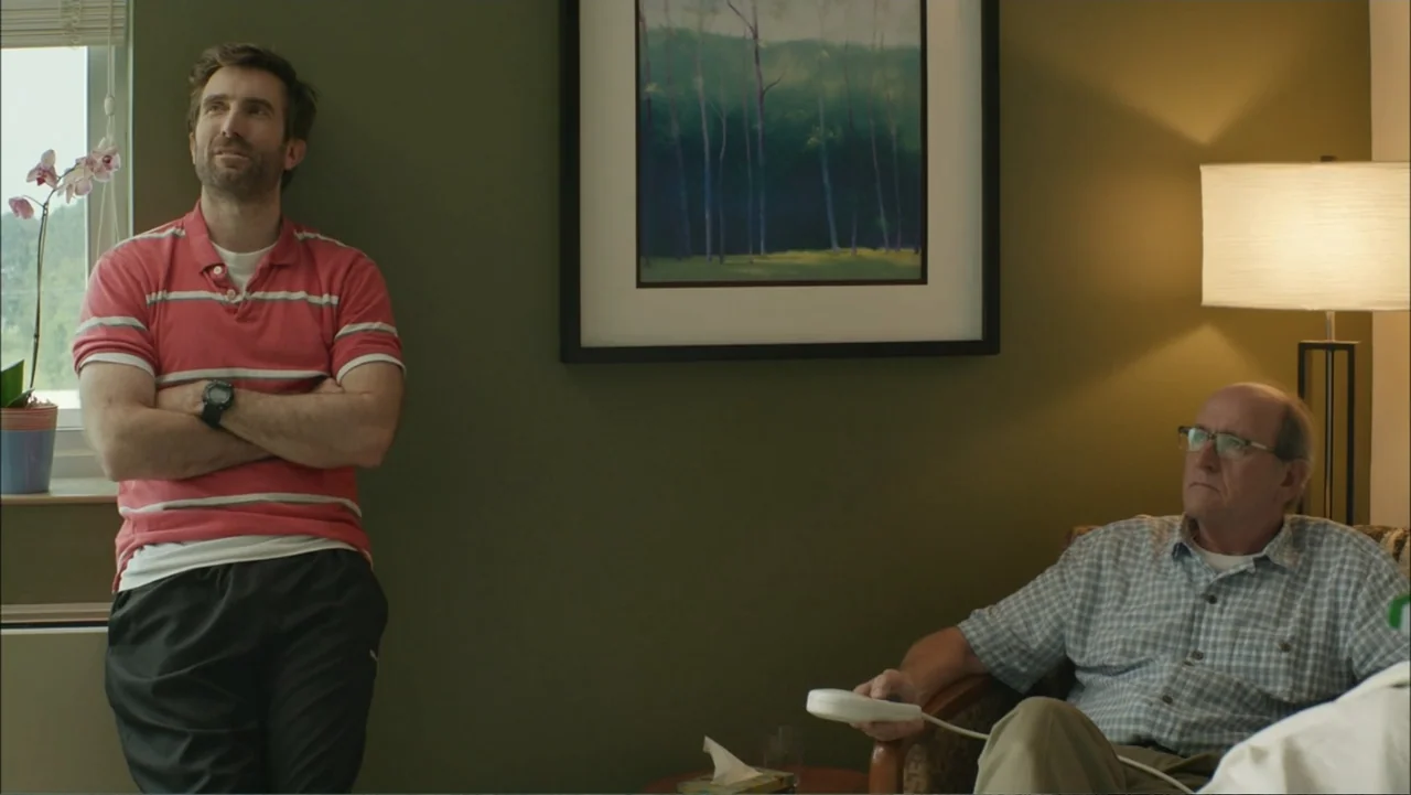 The Hollars (2016) movie still: medium — An interior room has dark olive walls and a framed landscape print hanging center. Two men stand and sit…