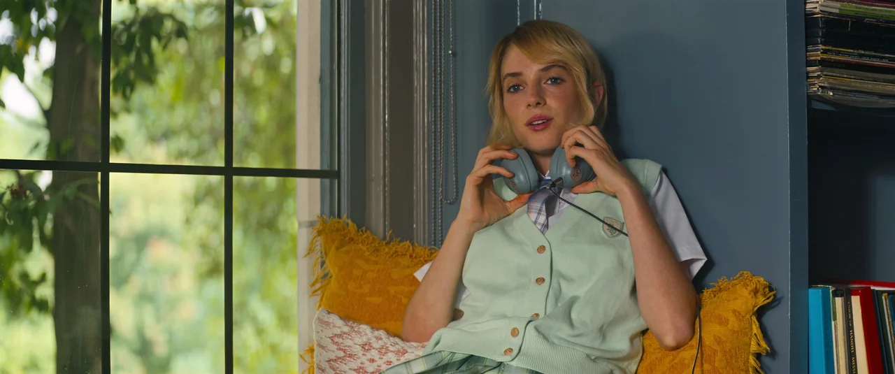 Do Revenge (2022) movie still: medium — A window seat features a young woman resting against a blue wall with yellow pillows. The subject holds…