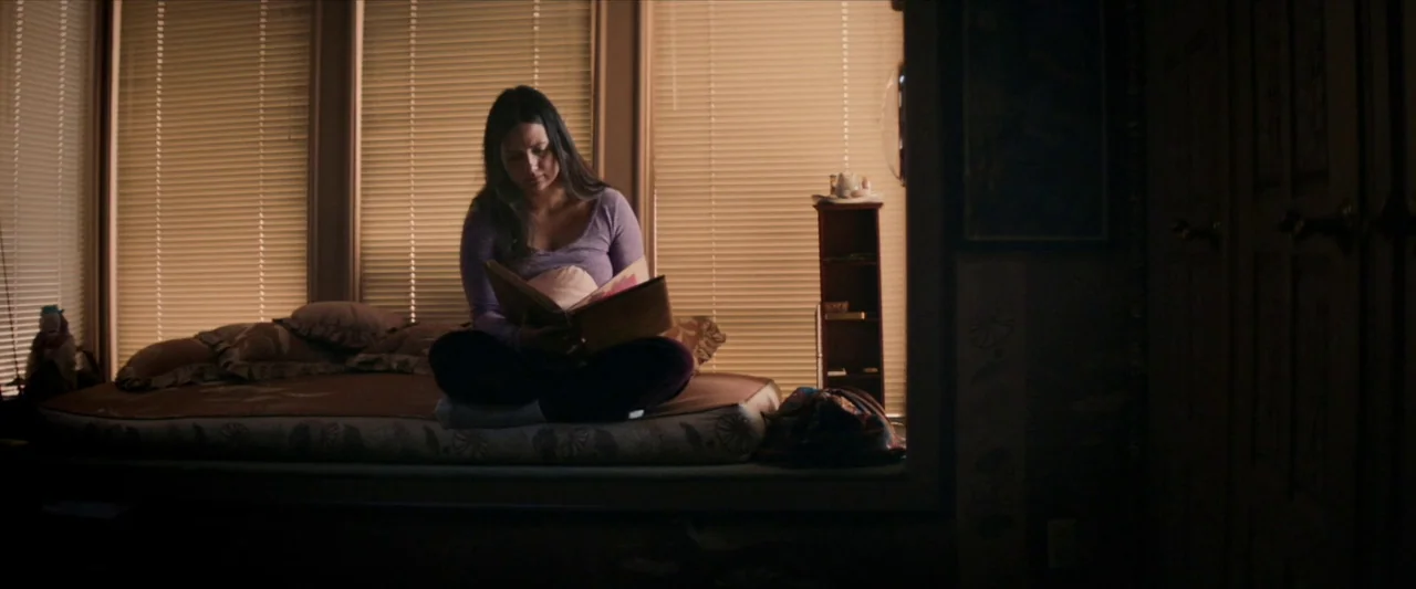 The Unknown Country (2022) movie still: medium — A woman sits on a window seat in front of closed horizontal blinds. She holds an open book and…