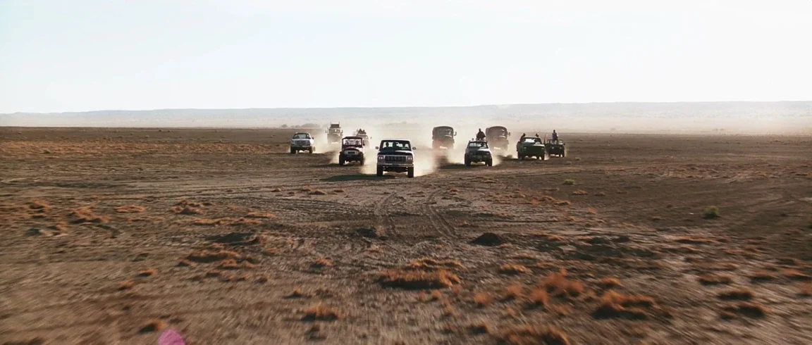 Transcendence (2014) movie still: wide — An expansive, arid desert plain extends to the horizon under a pale sky. A convoy of various SUVs and…