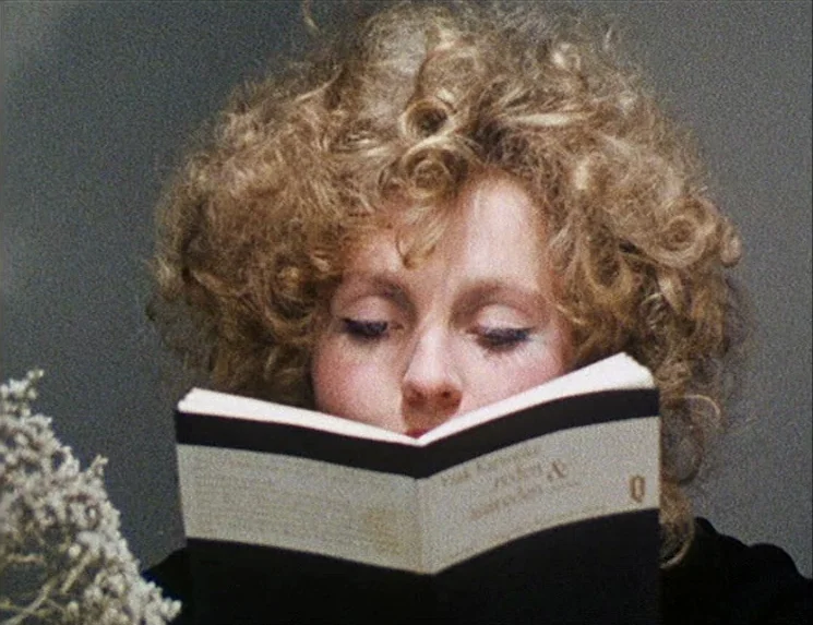 Rio Das Mortes (1971) movie still: closeup — A close-up shot of a young woman with curly hair reading a book. Her eyes are closed while she holds…