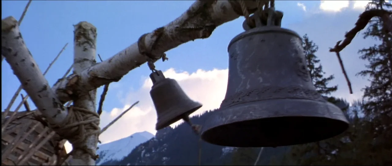 Ravenous (1999) movie still: closeup — Two metal bells are suspended from a rustic white birch wood support beam. In the distance, snow-capped…