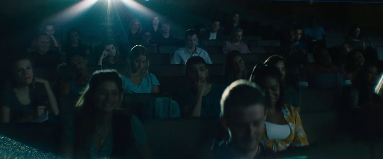 The Mad Hatter (2021) movie still: wide — A dimly lit tiered lecture hall filled with students looking forward toward a screen. A projector beam…