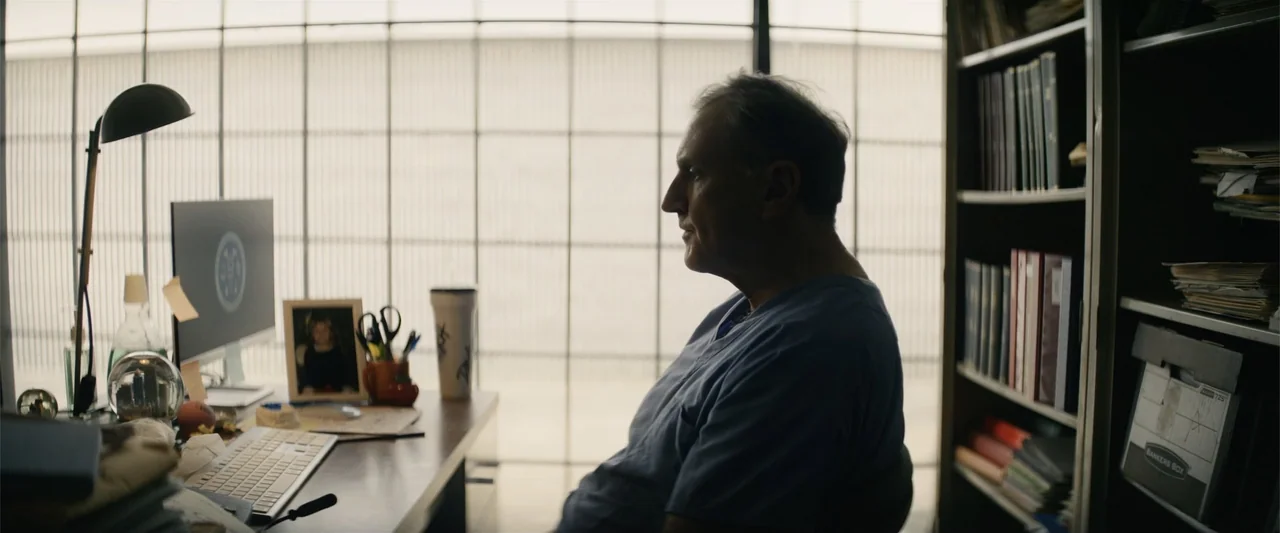 Last Looks (2022) movie still: medium — A cluttered office features a desk with a computer and a bookshelf. A man in medical scrubs sits at the desk…
