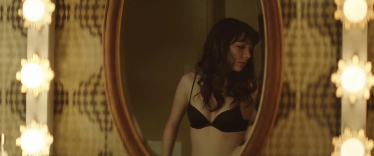 Southbound (2015) movie still: medium — A woman stands before a vanity mirror with patterned wallpaper in the background. She wears a black bra and…