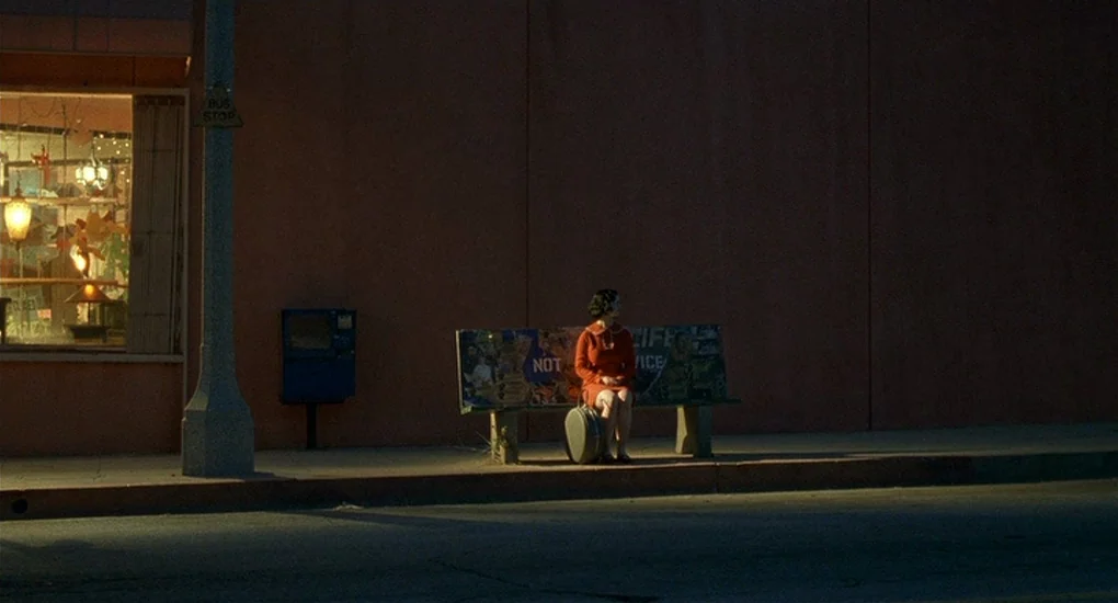 Ghost World (2001) movie still: A solitary figure in a vibrant red dress sits on a weathered bus stop bench at dusk, dwarfed by the expansive, dark…
