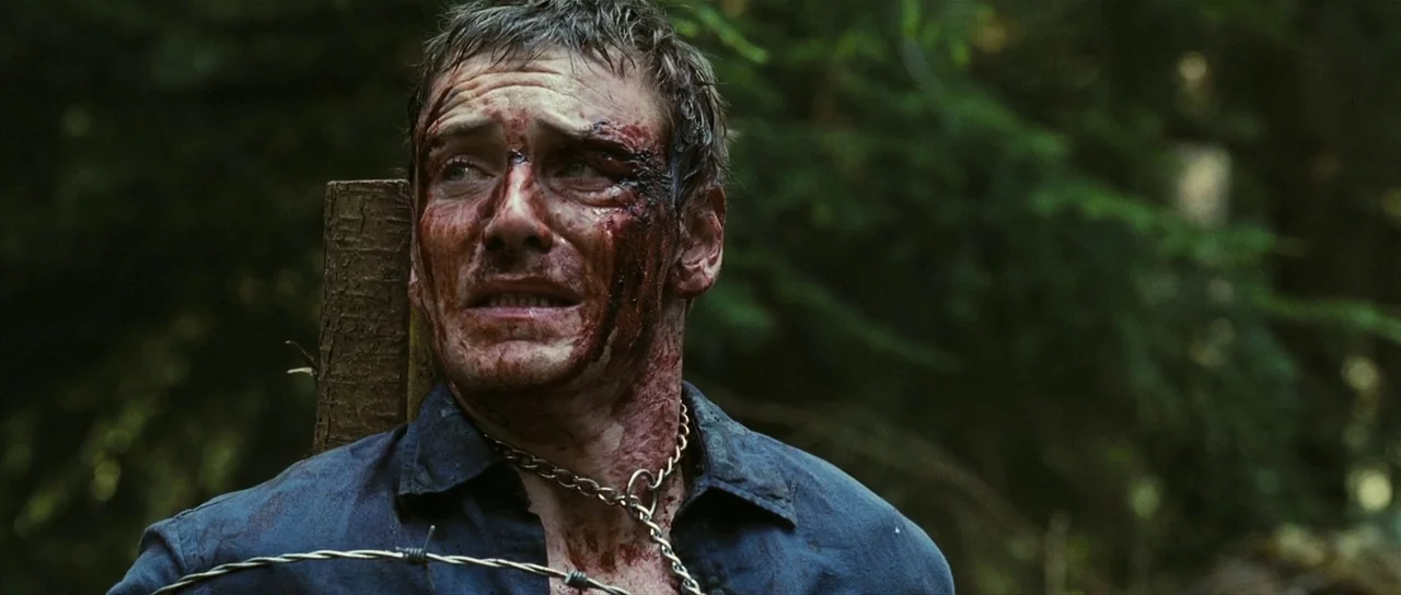 Eden Lake (2008) movie still: A visceral and intense closeup of a bloodied man restrained against a wooden post in a dense forest. The composition…