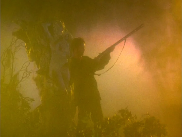 Twilight of the Ice Nymphs (1997) movie still: fullbody — A man stands in a dense, yellow-hazed outdoor space near a large stone statue. He holds a…