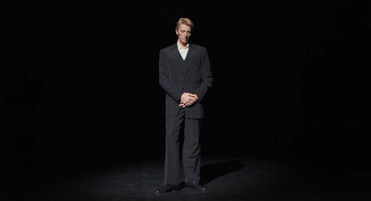 Pina (2011) movie still: fullbody — An empty dark void serves as a minimalist stage for a single person. A man in a pinstriped suit stands alone…