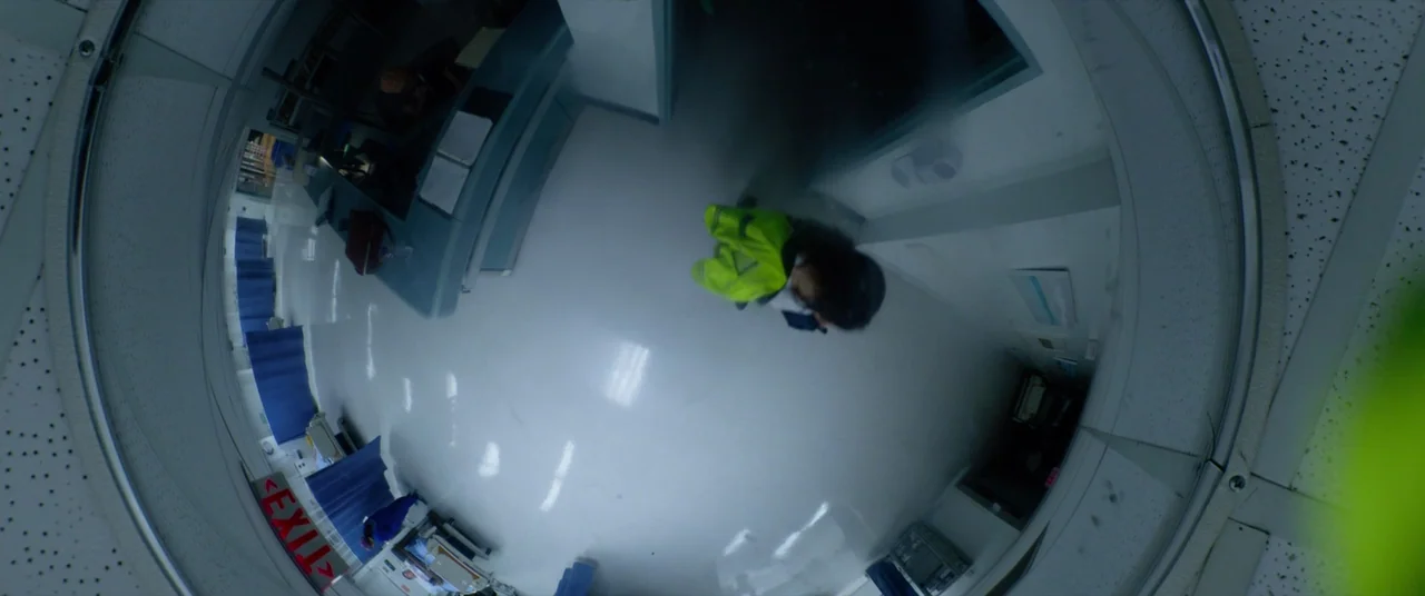 Ambulance (2022) movie still: wide — A top-down view of a sterile medical facility with white tile floors and blue curtains. A person in a…