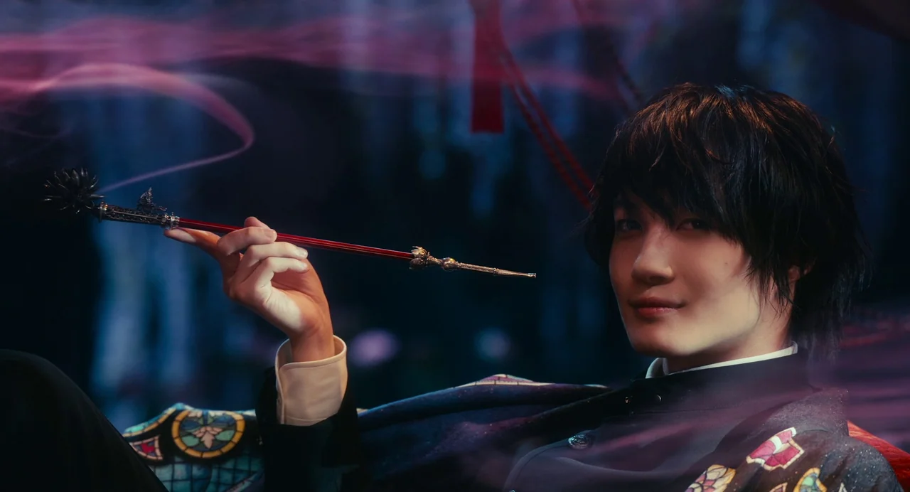 xxxHolic (2022) movie still: closeup — A dark, stylized interior features subtle colorful light refractions in the background. A young man holds a…