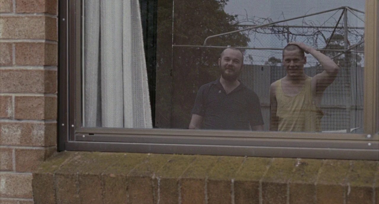 Snowtown (2011) movie still: medium — A view through a window reveals two men standing in an interior space. The exterior features a brown brick…