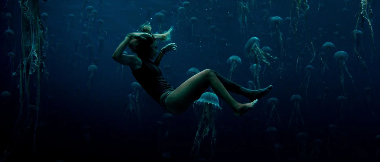 The Shallows (2016) movie still: fullbody — A woman drifts through deep blue water densely populated by translucent glowing jellyfish. The subject…