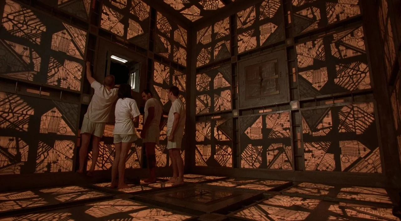 Cube (1997) movie still: wide — A square room features metallic walls covered in repeating geometric circuit-like patterns glowing orange. Four…
