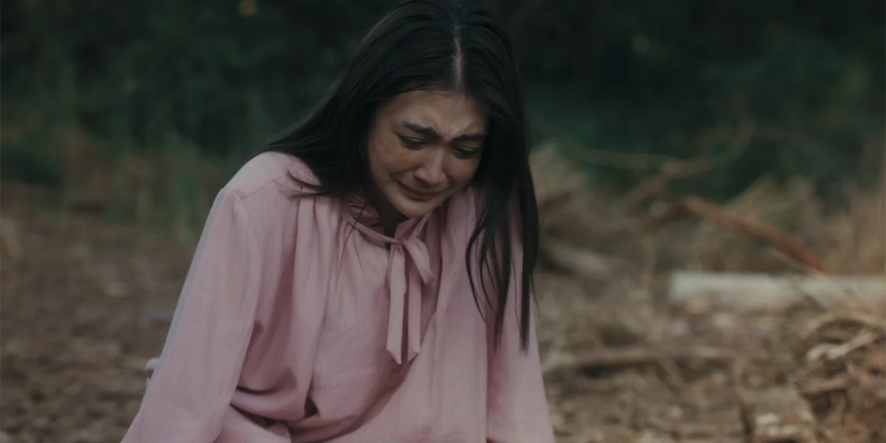 Host (2020) movie still: medium — A woman kneels on dry brown earth covered with small branches and twigs. The woman wears a pink blouse and cries…