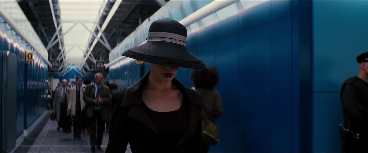 The Dark Knight Rises (2012) movie still: medium — A long, bright blue airport walkway features travelers walking in the background. A woman in a…