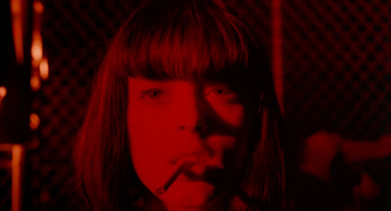 The Doom Generation (1995) movie still: closeup — A dimly lit room is bathed in deep monochromatic red light. A young woman with dark hair stares…