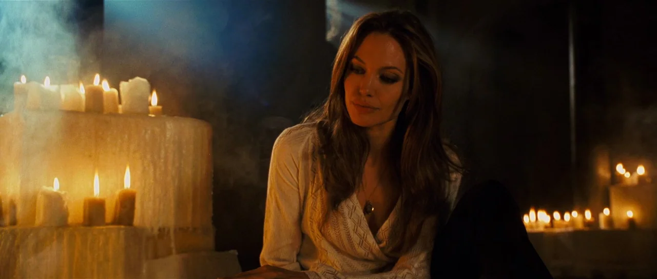 Wanted (2008) movie still: medium — A dark, dimly lit stone chamber is filled with clusters of wax candles. A woman with long wavy brown hair sits…