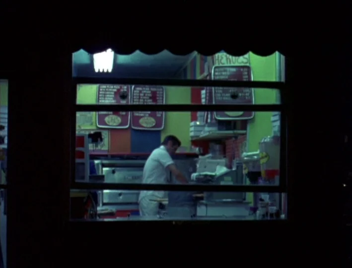 News From Home (1977) movie still: medium — A small storefront interior at night features bright lime-green walls and dark menu boards. A man in a…