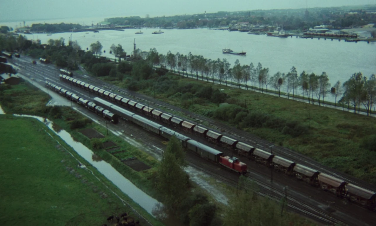 Wrong Move (1975) movie still: establishing — An overhead view shows a long freight train traveling along tracks beside a river. Green fields,…