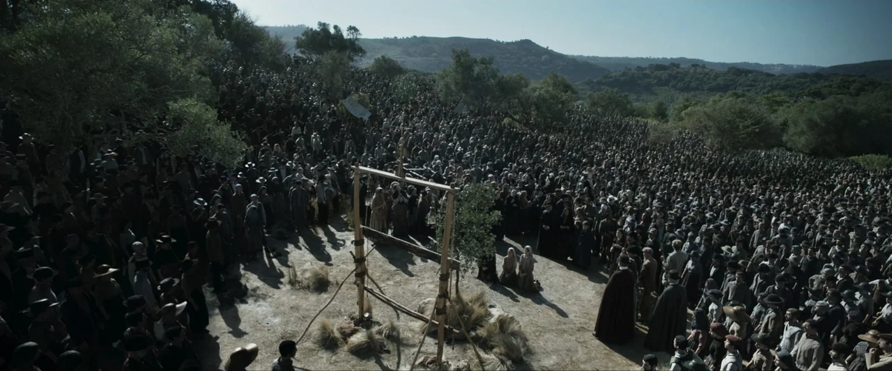Fatima (2020) movie still: wide — A vast crowd gathers on a hillside in a barren, dusty rural landscape. Two women kneel beneath a primitive wooden…