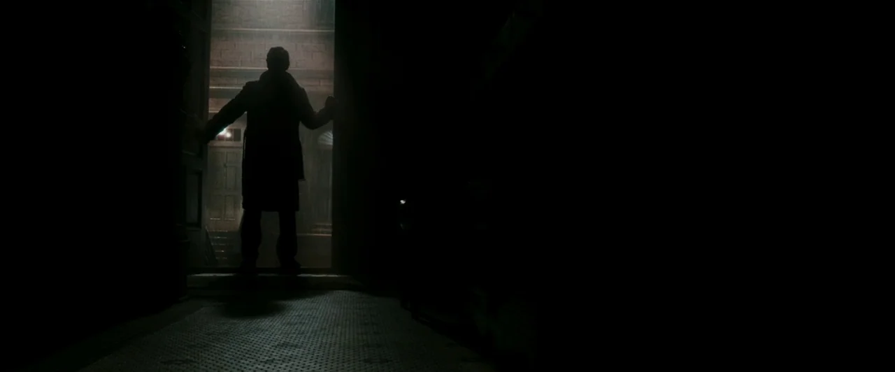 Watchmen (2009) movie still: wide — A dark hallway leads to an open exterior door with light spilling inward. A man stands in the doorway looking…