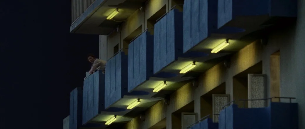 Munich (2005) movie still: wide — A modern multi-story apartment block with blue balconies at night. A lone man stands on the top balcony looking…