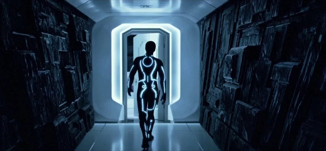 TRON: Legacy (2010) movie still: fullbody — A narrow corridor features dark, intricately textured geometric walls leading to an illuminated white…