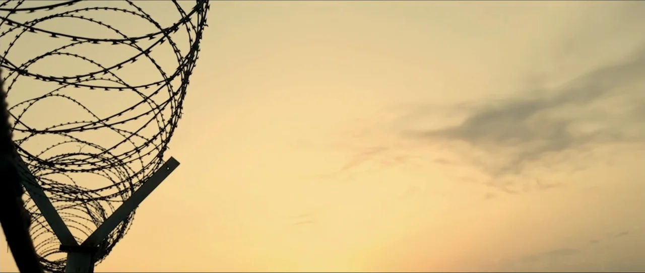 American Sicario (2021) movie still: establishing — A tall perimeter fence topped with coiled barbed wire stands against a clear sky. The setting…