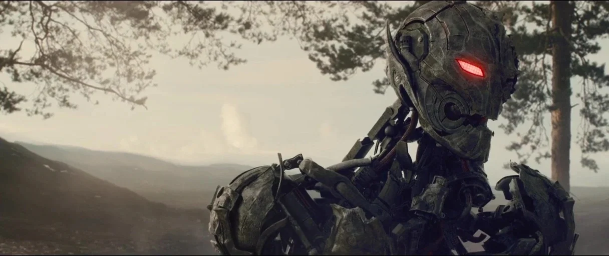 Avengers: Age of Ultron (2015) movie still: medium — A metallic humanoid figure stands in a wooded area with distant rolling hills visible. The…