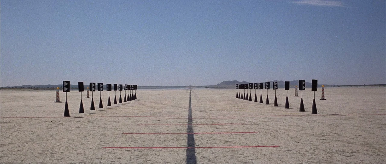 The Adventures of Buckaroo Banzai Across the 8th Dimension (1984) movie still: establishing — A vast, cracked dry lake bed features two rows of…