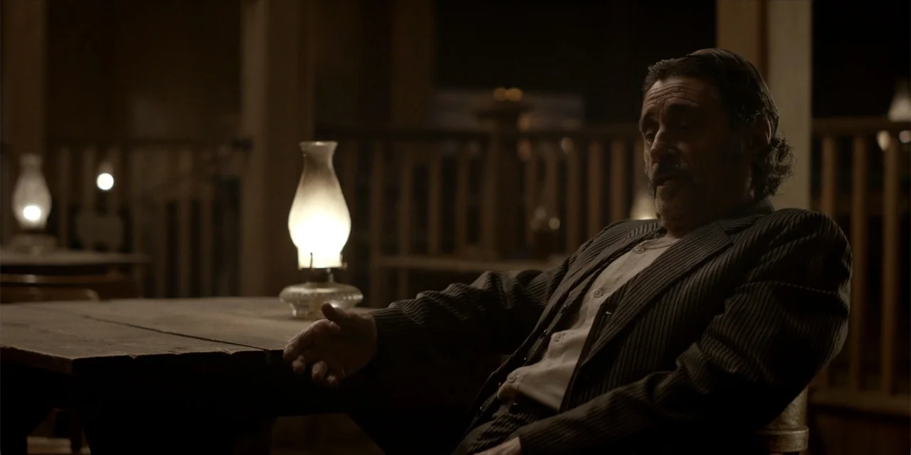 Deadwood: The Movie (2019) movie still: medium — A dark wooden saloon interior at night features tables illuminated by small oil lamps. A…