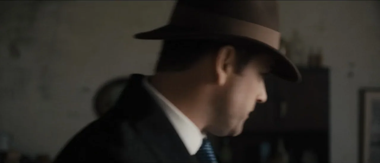 Fear Below (2025) movie still: closeup — A dimly lit room with plain walls and dark furniture in the background. A man wearing a fedora and suit…