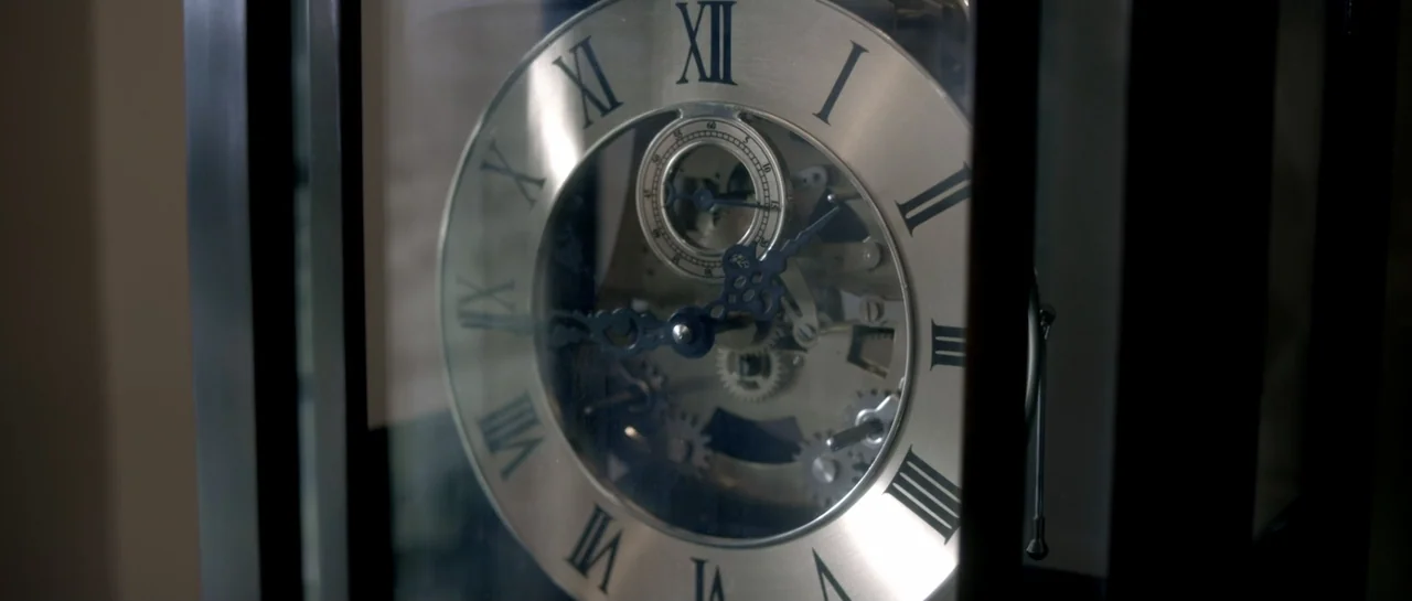 The Lullaby (2019) movie still: closeup — A close view of a metallic clock face with Roman numerals behind glass. The internal gears are visible…