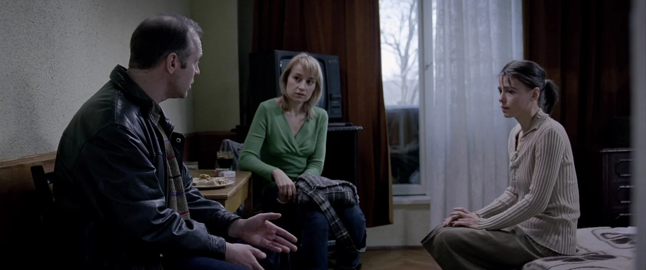 4 months, 3 Weeks and 2 Days (2007) movie still: medium — A modest apartment interior with plain walls and dark curtains by a window. Three people…