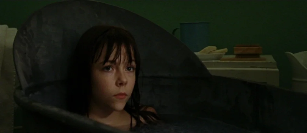 Innocence (2005) movie still: closeup — A dark room with a green-walled backdrop features a vintage metal bathtub. A young girl with wet hair sits…