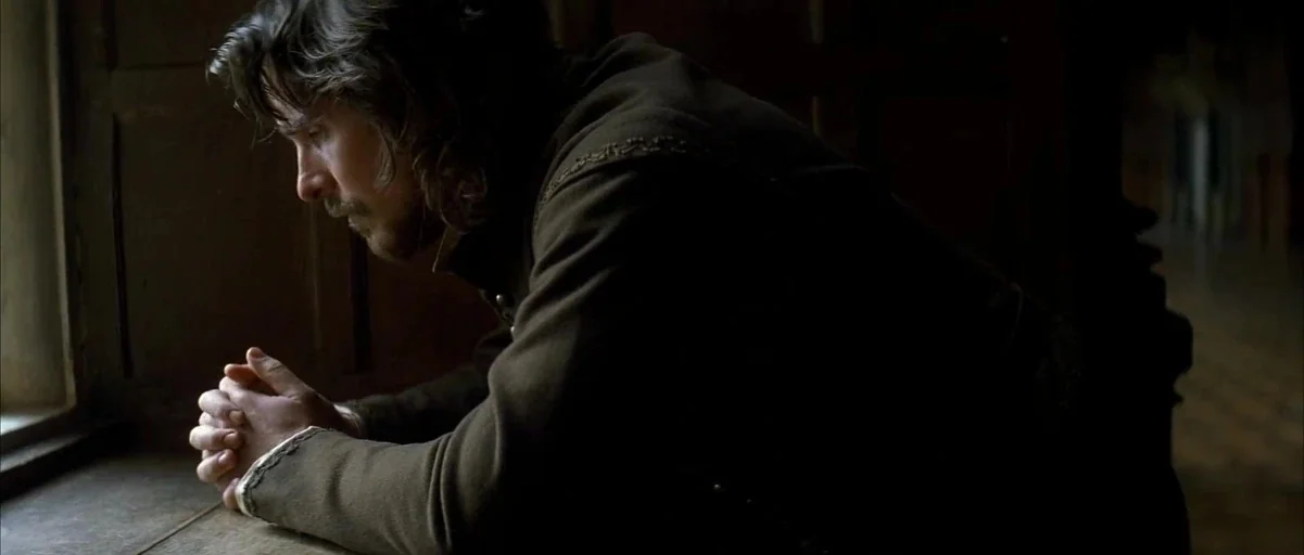 The New World (2005) movie still: medium — A dark, wood-paneled room interior with light entering from a side window. A man stands in profile,…