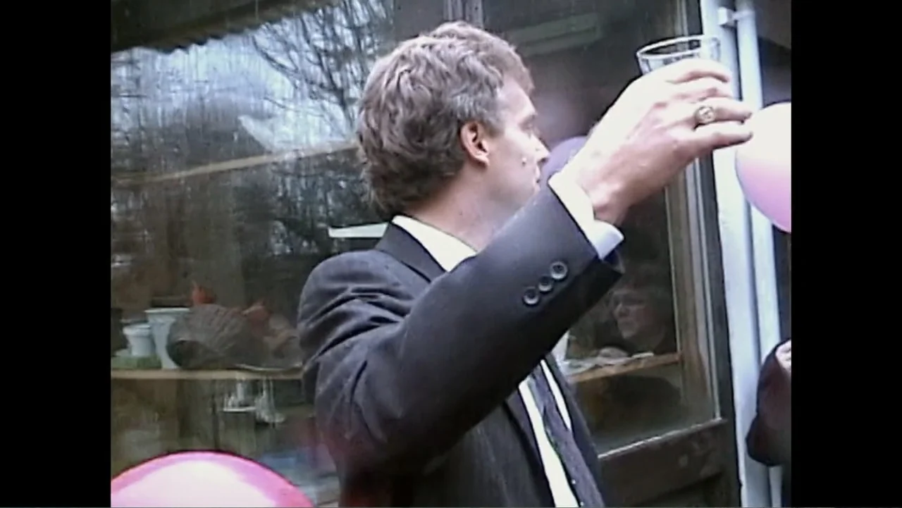 Daniel (1983) movie still: medium — A person stands outdoors near a windowed structure during the day. A man in a suit raises a glass in an upward…