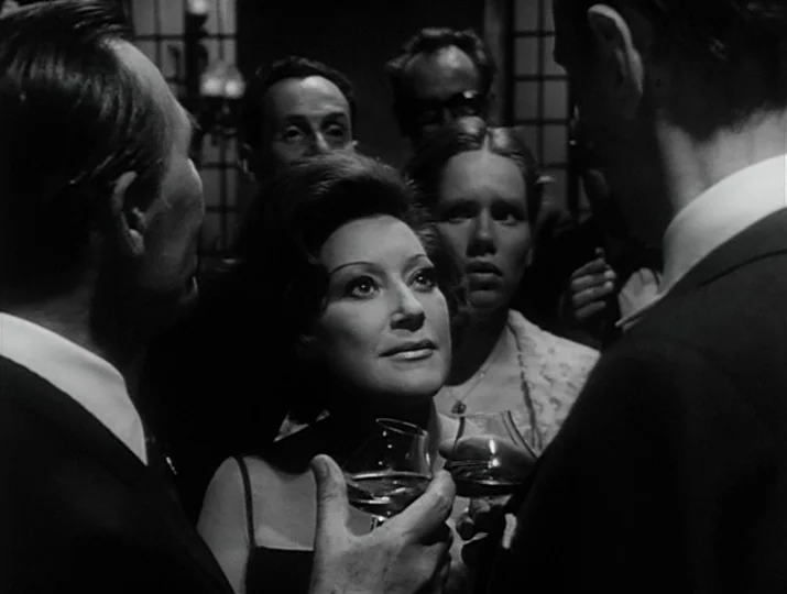 Hour of the Wolf (1968) movie still: medium — People socialize during an evening event, gathered closely in a dimly lit indoor space. A woman stands…
