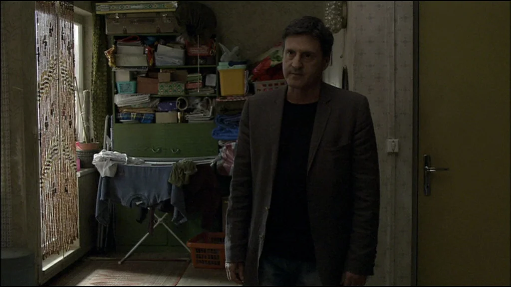 Caché (Hidden) (2005) movie still: medium — A cluttered apartment room features a green cabinet, a drying rack, and stacked boxes. A middle-aged man…