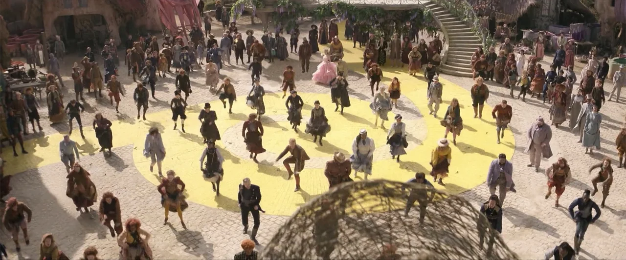 Wicked (1998) movie still: wide — A spacious stone-paved town square features a large swirling yellow painted path. A dense crowd of people in…