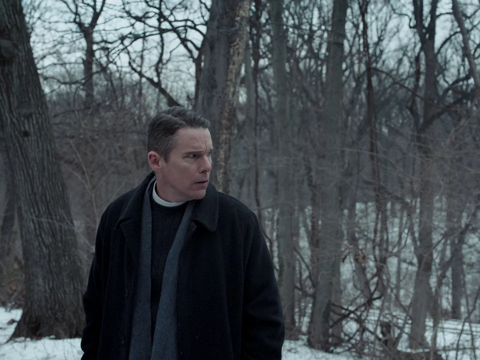 First Reformed (2017) movie still: medium — A dense patch of leafless trees stands in a snowy, overcast woodland environment. A man dressed in…