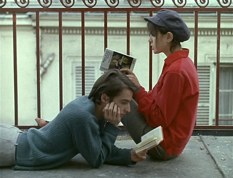 La Chinoise (1967) movie still: fullbody — Two individuals sit on a rooftop balcony enclosed by a wrought iron railing. The man reclines on the…